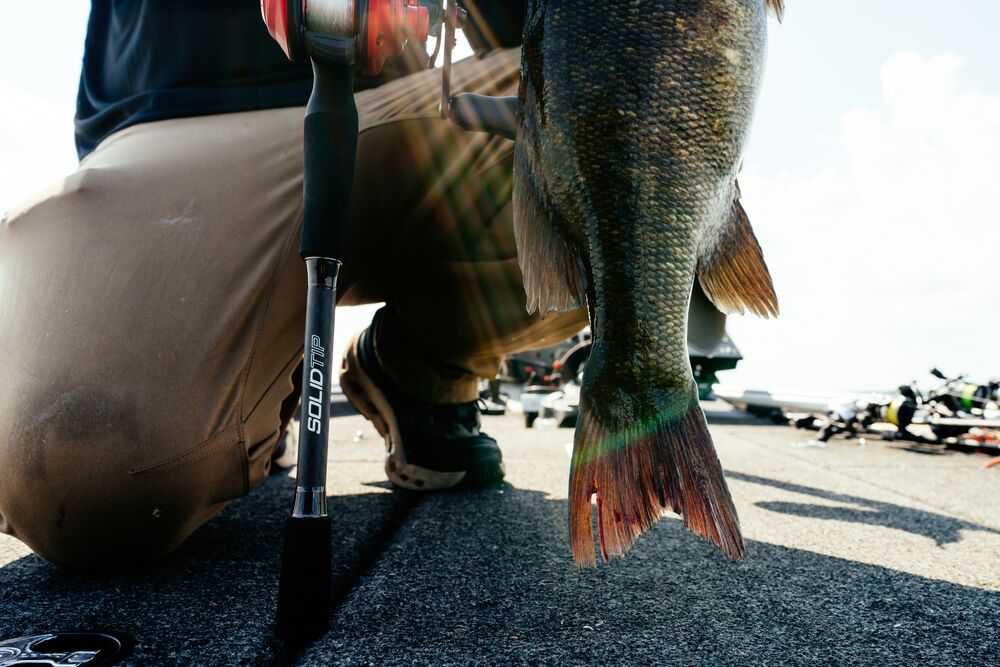 A close-up action shot of a person kneeling on a boat deck holding a freshly caught largemouth bass, with a Abu Garcia Veritas Tournament Solid Top Rod standing nearby.