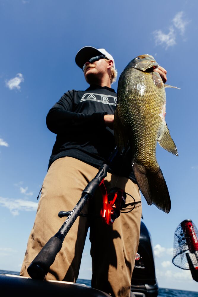 Man wearing Abu Garcia branded black sweatshirt and khaki pants holding a large fish with the Abu Garcia Veritas Tournament Rod leaning against him.  