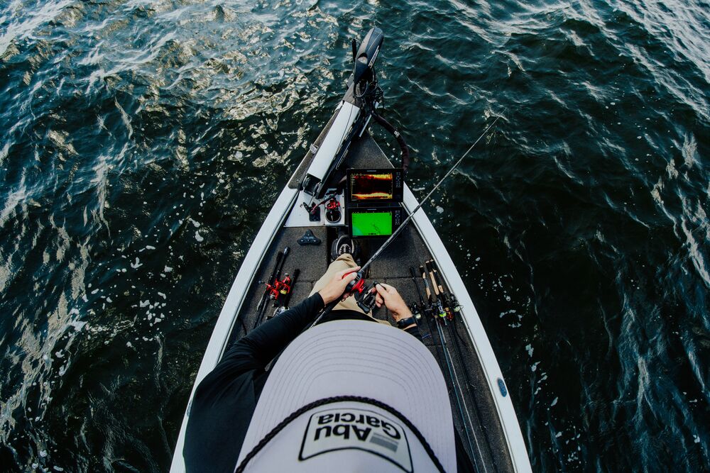 Overhead view of a person on a fishing boat holding a Abu Garcia Veritas Tournament Solid Tip rod, surrounded by dark blue water. The person wears a white and black shirt with the brand name 'Abu Garcia' visible on the hat. 